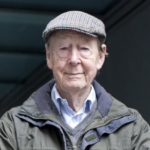 Sir Tom Kibble, dressed in a newsboy cap, a blue shirt layered with a grey sweater and a green jacket.